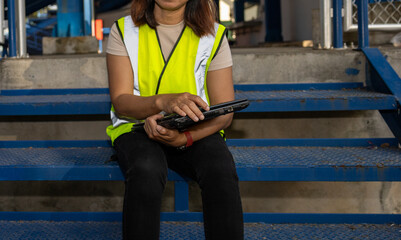 A worker in a safety vest uses a laptop while sitting on blue metal steps in a busy industrial site during the day. Machinery is in the background