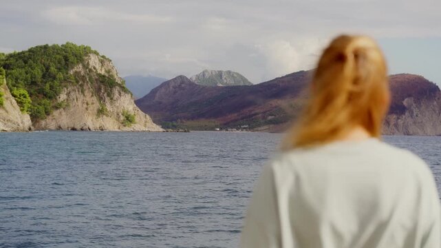 A blurred woman on a foreground stands by the water in Petrovac, Montenegro watching the mountains in the distance
