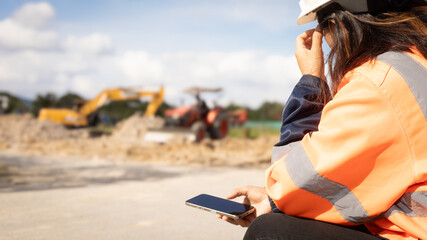 A worker in safety gear sits on a construction site looking at their phone while heavy machinery operates in the background. Activity is ongoing during the day