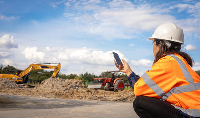 Construction worker in safety gear squats while looking at a phone. Heavy machinery like a tractor and excavator is at work in the background under a clear sky © Happy Photo