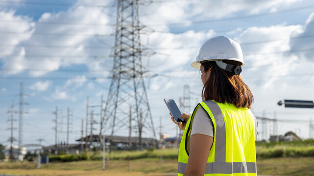 A worker in a safety vest and helmet stands outside, looking at a mobile device. Power lines and a tall structure background indicate a construction site during the day
