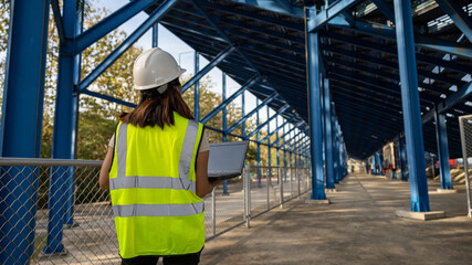 A worker stands at a construction site wearing a hard hat and a safety vest. She looks at a laptop while being surrounded by metal structures and clear skies © Happy Photo