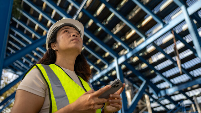 A worker wearing a hard hat and safety vest looks up at metal beams at a construction site. The worker holds a phone in hand while observing surroundings