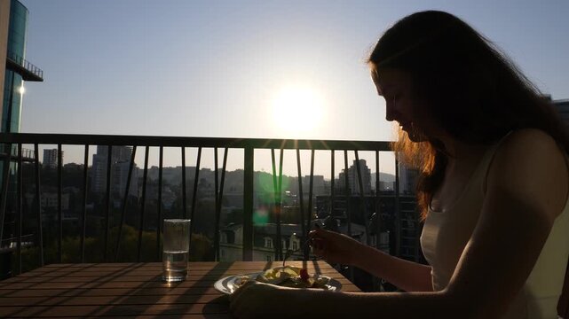 Young woman sit at sunny morning on apartments balcony with fork in salad plate, unhappy with healthy breakfast. Capricious girl look to lettuce leaves and finick with fork