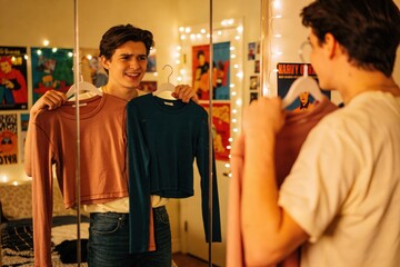 Young smiling man holds up two fashionable crop-top shirts, comparing his outfit options in the reflection of his bedroom mirror