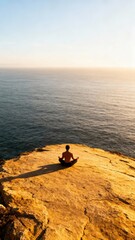 A fit young man is sitting in a meditative yoga pose on a high rocky cliff overlooking the vast ocean during sunset and copy space