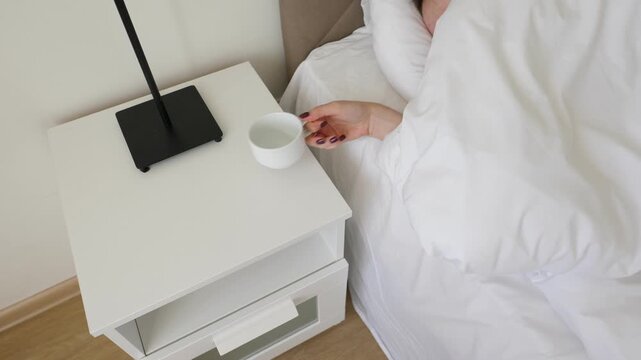 Woman hand take tea cup with pure water from bedside table and move under blanket. Sleepy girl dont want to wake up, have simple drink while remains laying on bed