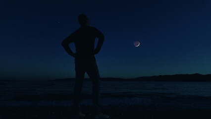 Man on the beach with young crescent Moon with stars and planets over ocean sea waters.