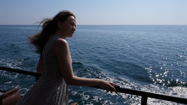 Dreamy woman enjoy short tour at sea, stand lean to old ship railings, long wavy hail fly on breeze. Girl musingly look ahead, tilt head, nice calm sea surface with shimmering ripples under sunlight.