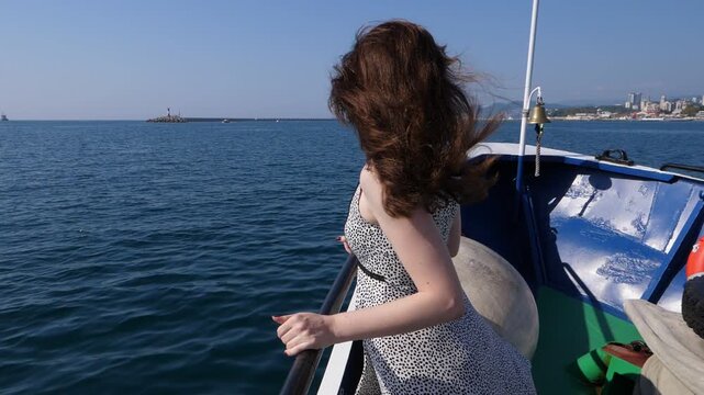 Young happy woman enjoy nice short tour at old ship, long wavy hair fly in wind, light dress skirt flutter down. Girl look ahead to port, calm sea water surface, slow motion shot.