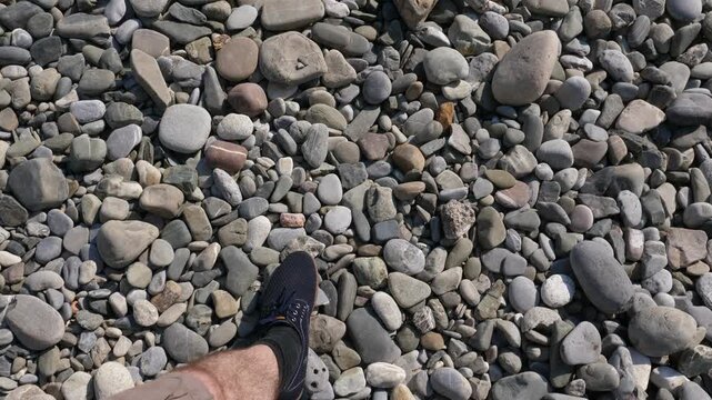 Man in light shoes walk by shingle beach, top-down POV shot. Small rounded stones, nice looking natural surface at sea shore. Dry pebble particles under hot sun light