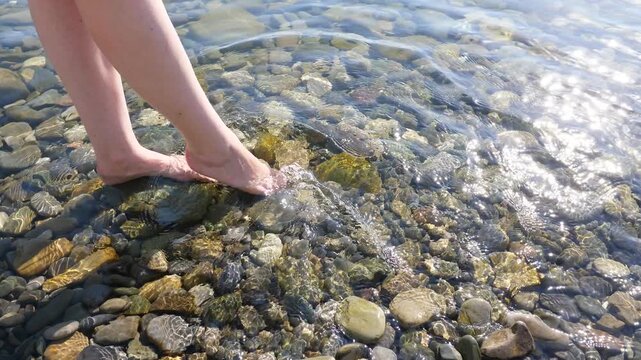 Young adult woman enjoy warm sea, make water splashes by feet, nice pebblestone beach at Sochi. Crystal clear seawater, bright sparkling under sun light. Playful girl dangle leg to make swash