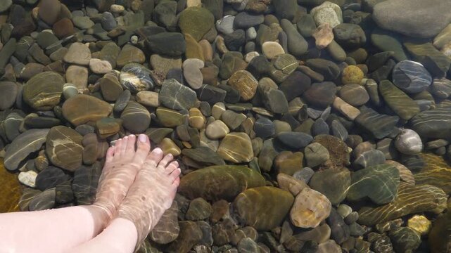 Young adult woman stand on rounded pebble stones at shallow place, crystal sea water, white skinned feets, POV style shot. Girl enjoy unusual beach, shingle at Black Sea shore