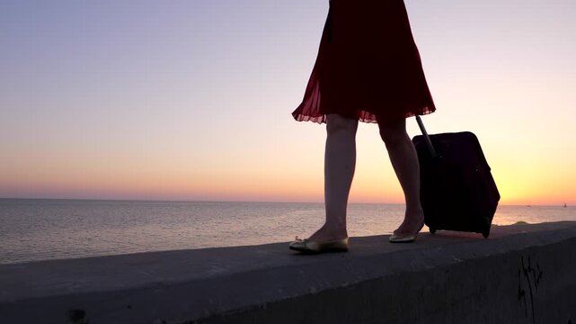 Slender young woman legs and trolley case, walking at embankment parapet against sunset. Traveller girl enjoy wonderful last evening at seaside, stroll with light luggage at dusk promenade.