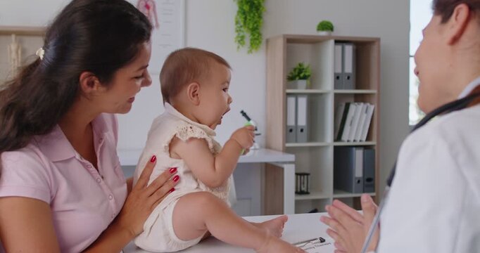 Mother holds baby on table as a pediatrician and family doctor prompt play with a green toy during a clinic appointment and checkup. Infant explores toy during supportive healthcare consultation.
