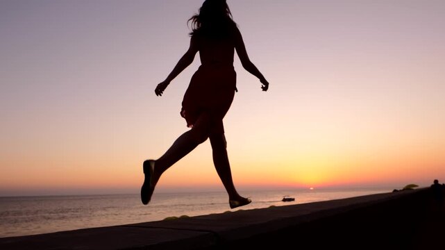 Young cheerful woman run at embankment parapet in high spirits, silhouetted shot against neat sunset sky and calm sea. Low camera move slower, girl rush and become blurred.