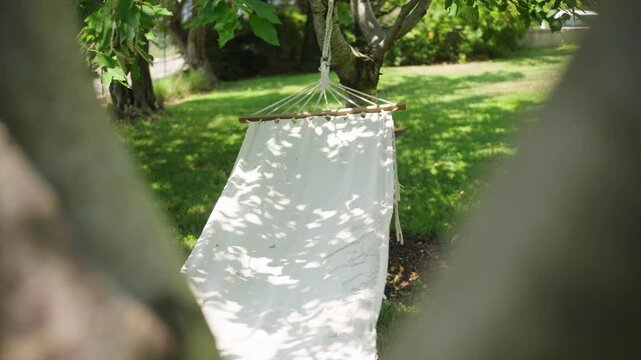 A white canvas hammock with a wooden spreader hangs between trees on a sunlit lawn. Soft bokeh and shallow depth of field emphasize fabric and rope details.