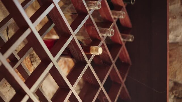 A wooden diamond wine rack holds corked empty bottles on a rough stone wall. Warm low key light, shallow depth, diagonal framing, slow pan and tilt reveal dust and cobwebs.