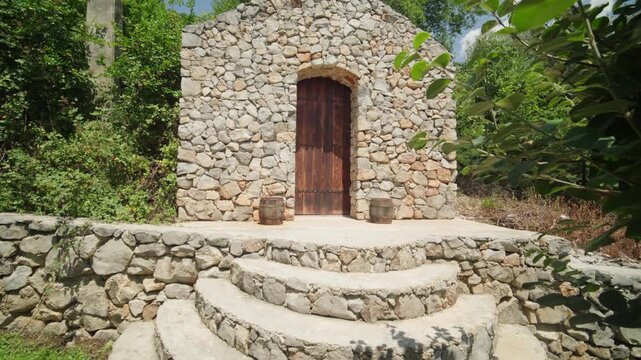 Rustic stone building with arched wooden door and semicircular tiered steps, two barrels and dense foliage. Camera slowly pushes in at midday, centered composition.