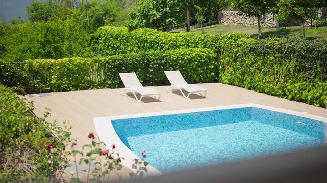 Sunlit private pool with bright blue mosaic tiling, two white lounge chairs, ivy hedges, stone wall, and tree lined hillside as the camera pans across the deck and water.