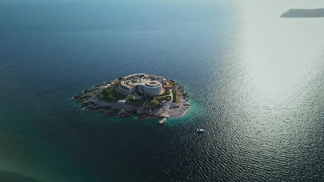 Aerial sequence shows Mamula Fort on Lastavica as a boat approaches the pier. Late afternoon glare cuts across deep blue sea and turquoise shallows off Montenegro.