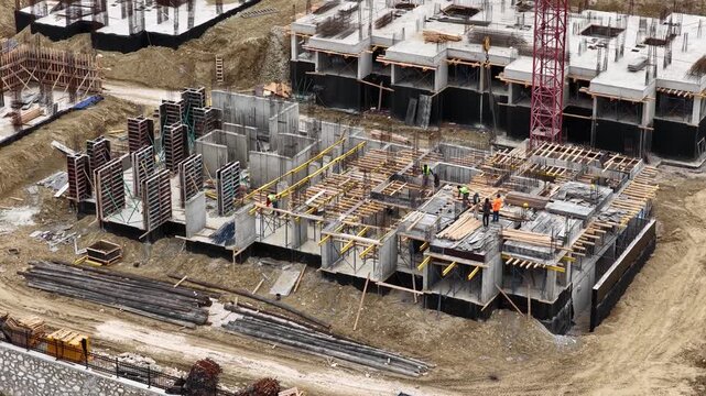 Aerial drone view of construction workers on a multi-story building site. Men in safety gear working on concrete formwork and reinforcement for a new residential project.