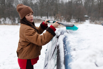 Woman driver removing snow from parked car in snowy area with trees in the background on clear cold day