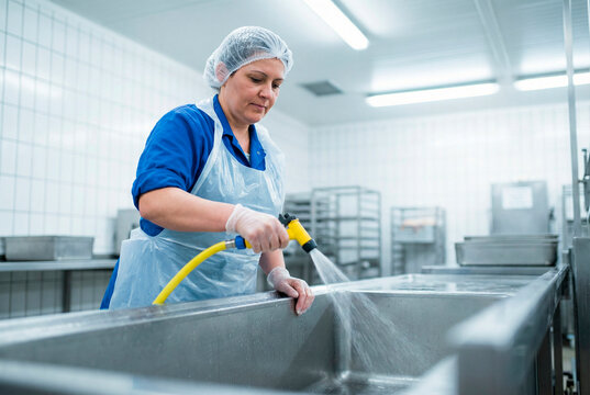 Female worker in protective gear cleaning a large metal sink with a yellow hose in a commercial kitchen setting with stainless steel equipment and white tiled walls.