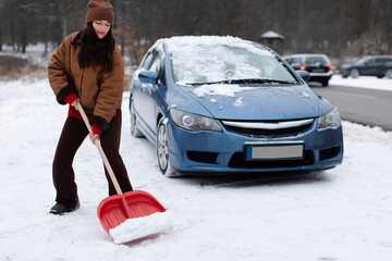 Woman clearing snow from the ground next to blue car in snowy area on winter day, full length shot