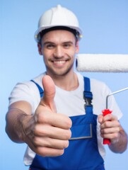 A cheerful painter in a hard hat and blue overalls shows a thumbs up while holding a paint roller. Home improvement and renovation concept.