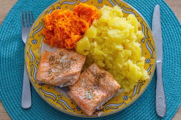 Zdrowy, domowy obiad składający się z pieczonego fileta z łososia, gotowanych ziemniak&oacute;w oraz sur&oacute;wki z tartej marchewki. Danie serwowane na dekoracyjnym ż&oacute;łtym talerzu ze sztućcami na niebieskiej pod