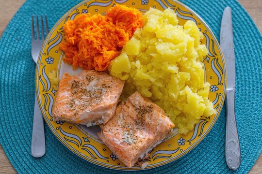 Zdrowy, domowy obiad składający się z pieczonego fileta z łososia, gotowanych ziemniak&oacute;w oraz sur&oacute;wki z tartej marchewki. Danie serwowane na dekoracyjnym ż&oacute;łtym talerzu ze sztućcami na niebieskiej pod