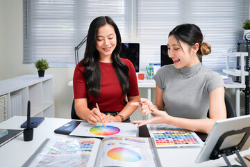 Two professional women collaborating on creative design project, discussing colorful color swatches, hues, and palettes at modern office workspace table. Ideal for wallpaper, poster