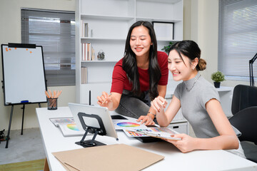 Two professional women collaborating on creative design project, discussing colorful color swatches, hues, and palettes at modern office workspace table. Ideal for wallpaper, poster