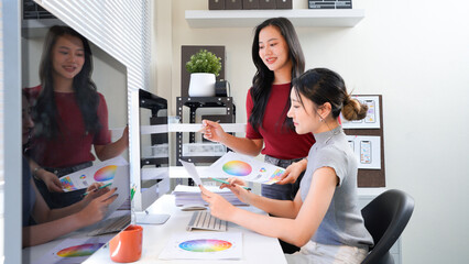 Two professional women collaborating on creative design project, discussing colorful color swatches, hues, and palettes at modern office workspace table. Ideal for wallpaper, poster