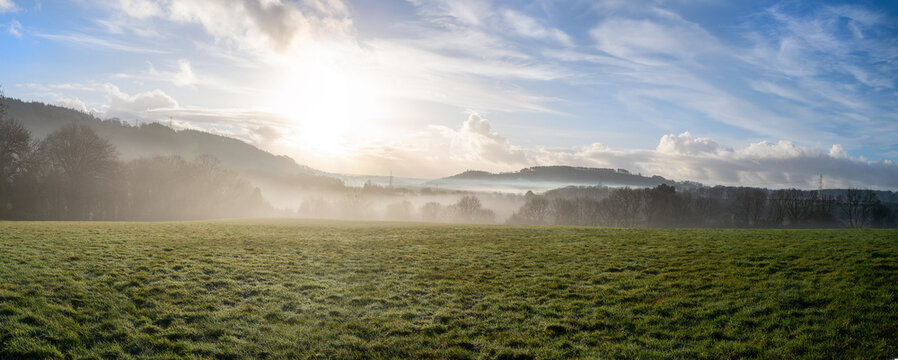 English countryside wide panoramic picture showing a green field at sunrise. Early morning mist and a vibrant blue sky add to the rural lifestyle and country life feel. Winter, autumn and seasons.