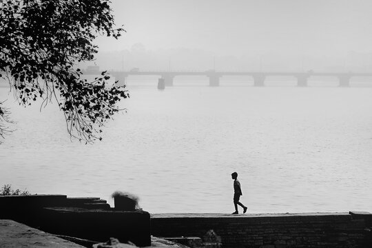 Lonely person walking alone along the river bank. Vast open space however bridge and a tree visible to connect with the social world