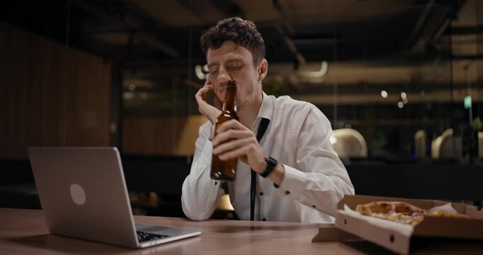 A young man takes a break in a quiet eatery, looking weary as he glances at his laptop, pizza, and drink nearby