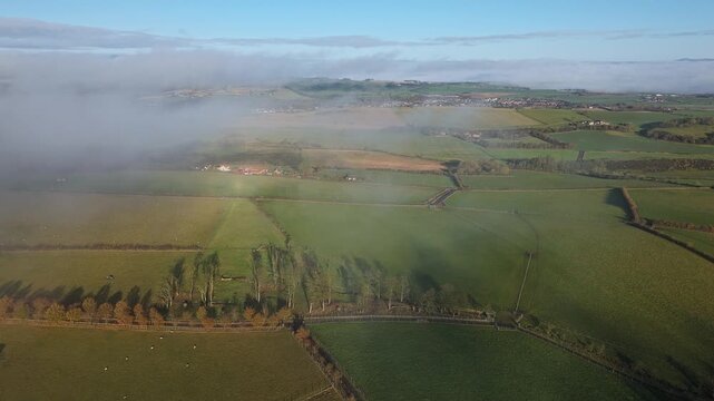 Drone footage of patchwork farmland and rural fields partially covered by drifting fog, revealing roads, trees and distant villages under clear blue sky.