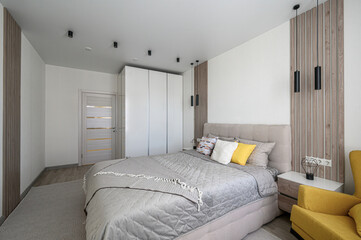 A modern bedroom with a beige upholstered bed, grey quilt, yellow armchair, wooden slat accent walls, white wardrobe, black pendant lights, and neutral rug