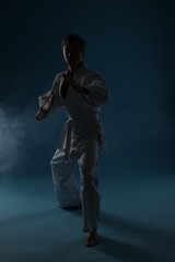 Man in uniform practicing karate on dark light blue background with smoke