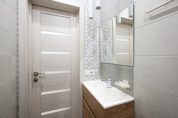 sleek modern bathroom with light grey tiles, wooden vanity with white countertop, chrome faucet, rectangular mirror, towel rack, and recessed lighting. Clean, minimalist design