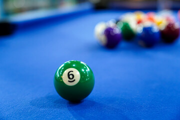 A green six billiard ball sits prominently on a blue felt pool table, ready for action. Behind it, a triangle of colorful billiard balls waits for a player to break in competitive.