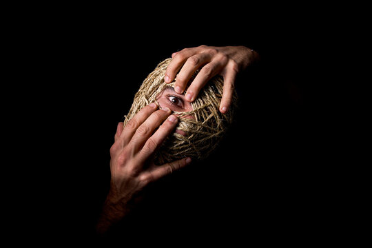 Conceptual portrait of a man with head wrapped in rope, showing one eye with an expression of anxiety, fear, and mental health struggle over a black background.