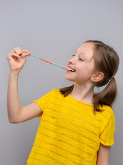 young girl in profile against a light gray background. girl is holding a long, thin, multicolored gummy candy stretched between her fingers and her mouth. 