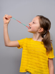 young girl in profile against a light gray background. She is wearing a bright yellow knitted shirt and has her light brown hair tied back in a ponytail. The girl is holding a long, multicolored gummy