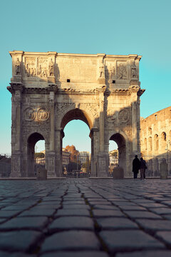 Low-angle view of the Arch of Constantine (Arco di Costantino) at golden hour in Rome, with warm sunset light highlighting the intricate marble carvings, reliefs, and triumphal arches. The ancient Rom