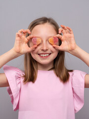 smiling young girl standing against a light gray studio background. She is wearing a soft pink dress and holding two heart-shaped, sugar-coated gummy candies over her eyes like playful glasses. The im