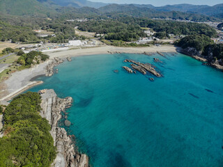 Fototapeta premium Wide aerial view of a turquoise coastal bay with rocky formations, sandy beach, and forested hills along the Shikoku coastline in Japan