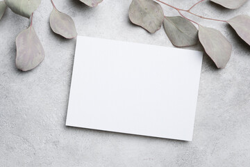 Blank white card with soft gray leaves arranged around it on a textured gray surface, ideal for invitations or greeting messages with copy space available